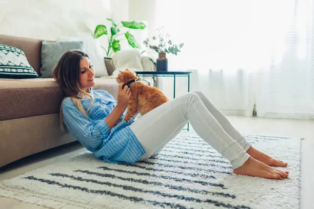 Photo of Playing with cat at home. Young woman sitting on carpet and hugging pet.