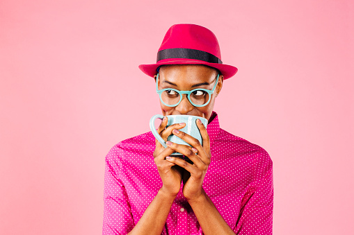 Portrait of a smart young woman with glasses drinking a coffee mug and looking to side, isolated on pink studio background