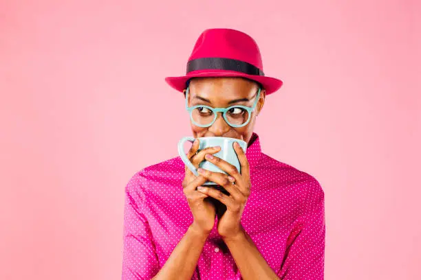 Photo of Portrait of a smart young woman with glasses drinking a coffee mug and looking to side, isolated on pink studio background