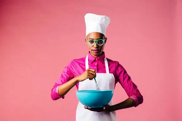 Photo of Portrait of a young woman in cooking hat and apron mixing a bowl with a whisker, isolated on pink studio background