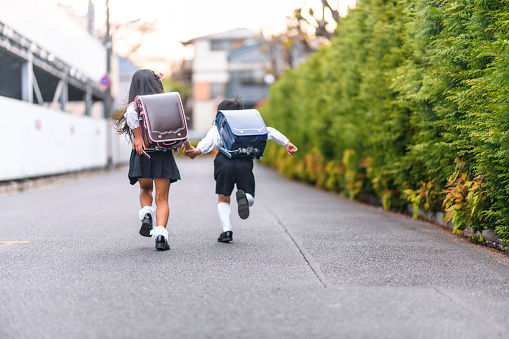 Rear view of two Japanese schoolchildren with randoseru holding hands and running along a pedestrian walkway.
