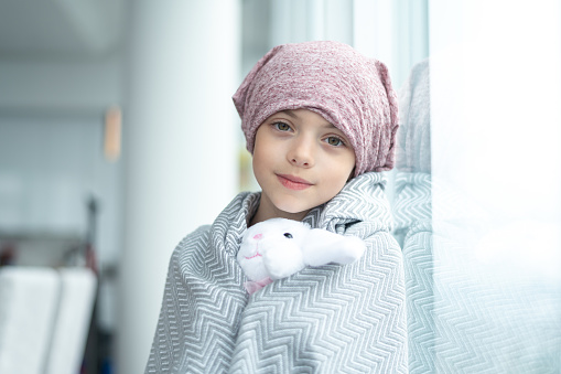 A happy elementary age girl with cancer is wearing a pink bandana on her head. She has a blanket draped over her shoulders and is holding a stuffed toy. The child is looking directly into the camera while leaning against a window. She has a thoughtful and serious expression.