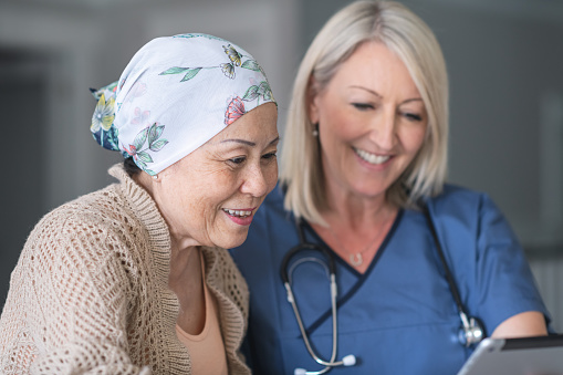 An female doctor is meeting with a mature adult patient fighting cancer. The patient is a Chinese woman. She is wearing a bandana to hide her hair loss from chemotherapy treatment. The two individuals are seated next to each other at a table.The physician is showing the patient test results on an electronic wireless tablet. Both women are smiling.