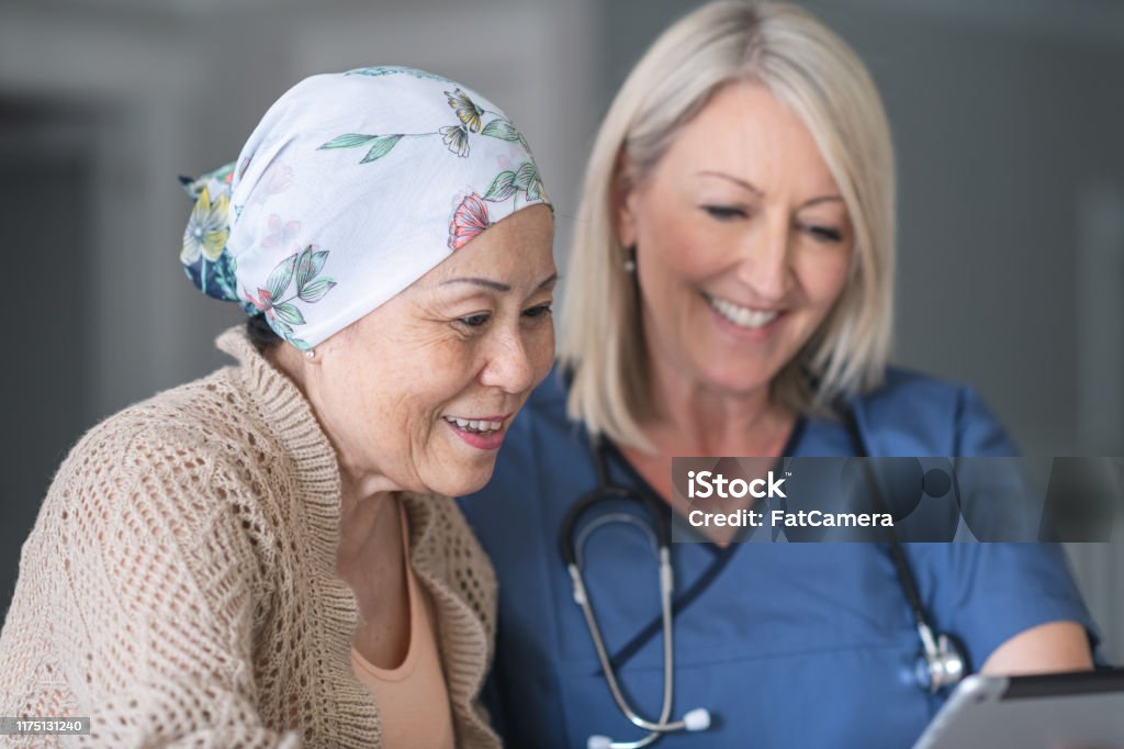 Female doctor consults patient with cancer An female doctor is meeting with a mature adult patient fighting cancer. The patient is a Chinese woman. She is wearing a bandana to hide her hair loss from chemotherapy treatment. The two individuals are seated next to each other at a table.The physician is showing the patient test results on an electronic wireless tablet. Both women are smiling. Cancer - Illness Stock Photo