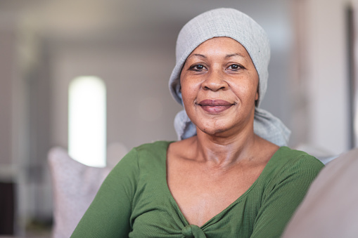 A mature black woman with cancer is wearing a scarf on her head. She is sitting at home in her living room. She is smiling directly at the camera. She is full of gratitude and hope for recovery.