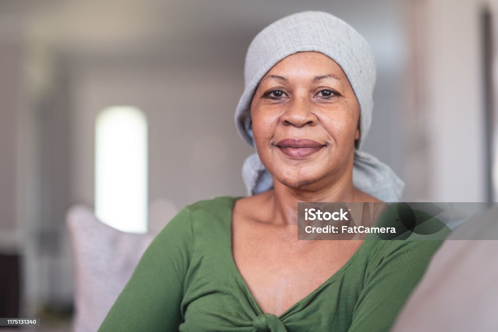 Portrait of a contemplative woman with cancer A mature black woman with cancer is wearing a scarf on her head. She is sitting at home in her living room. She is smiling directly at the camera. She is full of gratitude and hope for recovery. Cancer - Illness Stock Photo