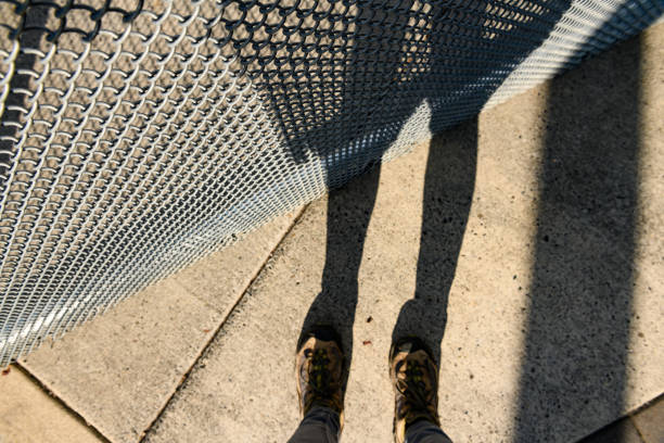 This is an abstract photograph of a woman’s shadow across a chainlink fence. She stands on the sidewalk in sneakers. Depth of field is shallow, focus is on the fence.