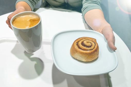 Female hands holding plate with cinnamon roll and white cup of coffee latte