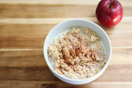 Apple Cinnamon oatmeal in a white bowl with milk. The bowl of oatmeal is served on a wooden cutting board with an fresh red apple.