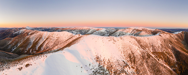 Wide aerial panorama of snowy mountain ridge on winter sunrise. Stunning mountains range covered with snow powder on ski resort at sunset. Caucasus mountain peaks skyline on a sunset.