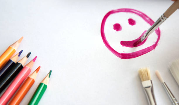 istock a child with a brush and raspberry paint draws a happy smile or smiling face on a sheet of white paper, on the sheet are colored pencils and brushes of different sizes. Place for text 1175166324