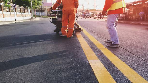 Road workers painting yellow lines on asphalt street Rear view of 2 road workers with thermoplastic spray marking machine working to paint yellow lines on asphalt road surface with flare light in evening time, selective focus road marking stock pictures, royalty-free photos & images