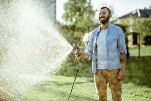 Handsome man watering green lawn, sprinkling water on the grass during a sunny morning on the backyard. Lawn care concept