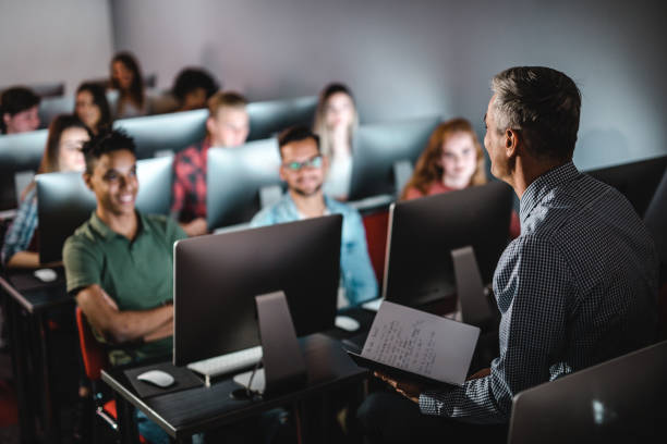 Mid adult male teacher teaching the lecture in computer lab. Male teacher talking to large group of students on a class at computer lab. computer lab stock pictures, royalty-free photos & images