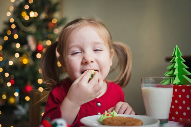 el niño come dulces en el fondo de un árbol de navidad con luces. cena de navidad de los niños - christmas child cookie table fotografías e imágenes de stock