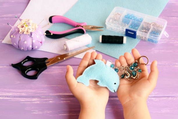 istock Child holds a dolphin in his hands. Home felt keychain with beads. Pincushion, thread, needles, pins, scissors, pliers, felt sheets, box of beads. Children summer crafts concept 1175183102