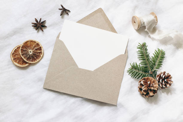 istock Christmas blank greeting card mock-up scene. Festive winter wedding composition. Craft envelope, pine cone, dry orange fruit slices and fir tree branch on white table, linen background. Flat lay, top view. 1175184108