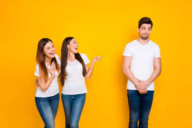 Portrait of excited girls mocking at guy standing, shy wearing white t-shirt denim jeans isolated over yellow background