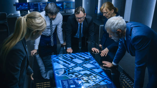 high angle shot of team of government intelligence agents talking while standing around digital touch screen table and tracking suspect. opération de surveillance par satellite dans la salle de surveillance. - protective suit photos photos et images de collection