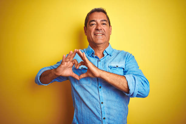 istock Handsome middle age man wearing denim shirt standing over isolated yellow background smiling in love showing heart symbol and shape with hands. Romantic concept. 1175201113