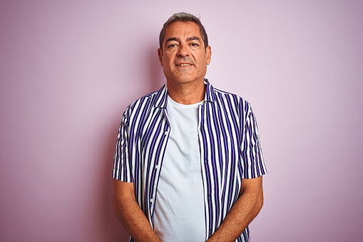 Handsome middle age man wearing striped shirt standing over isolated pink background with serious expression on face. Simple and natural looking at the camera.