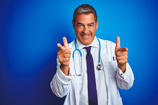 Handsome middle age doctor man wearing stethoscope over isolated blue background pointing fingers to camera with happy and funny face. Good energy and vibes.