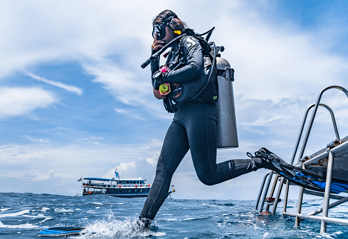 Andaman sea, Thailand - Aug 21 2019: One Asian Woman has jumped off a boat into the Andaman sea.  A scuba diver demonstrating perfectly what is known as the ‘giant stride entry’.  Regulator in her mouth and scuba equipment in place, she holds her mask in place.  Representative of the Millennial generation of eco tourists who spend much of their finances on environmentally friendly activities and travel.