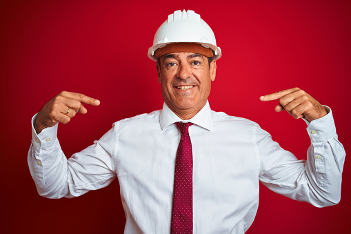 Handsome middle age engineer man wearing security helmet over isolated red background looking confident with smile on face, pointing oneself with fingers proud and happy.