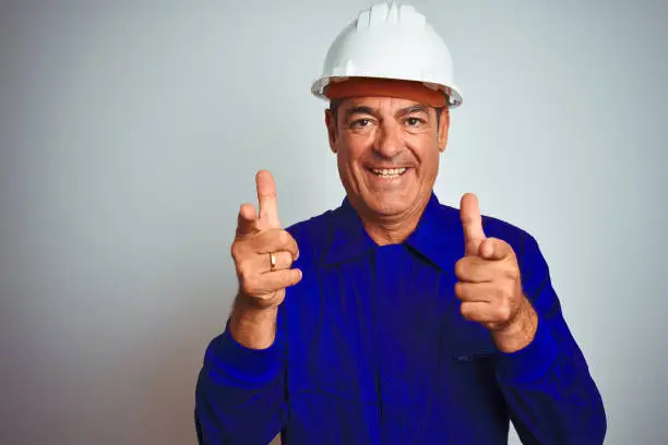 Photo of Handsome middle age worker man wearing uniform and helmet over isolated white background pointing fingers to camera with happy and funny face. Good energy and vibes.
