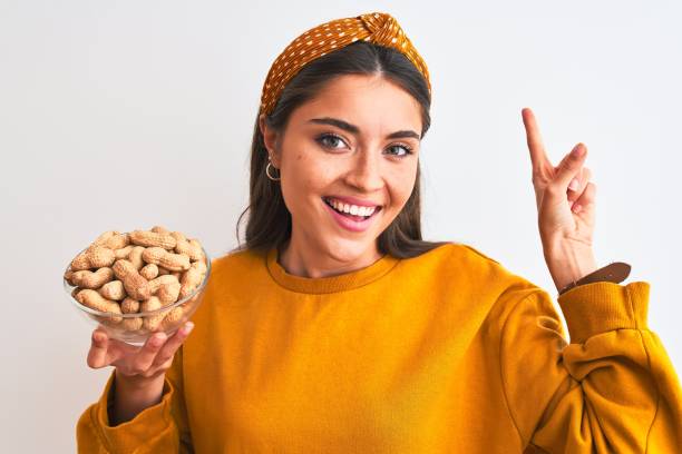 junge schöne frau hält schale mit erdnüssen stehen über isolierten weißen hintergrund sehr glücklich zeigt mit hand und finger zur seite - peanut food snack healthy eating stock-fotos und bilder