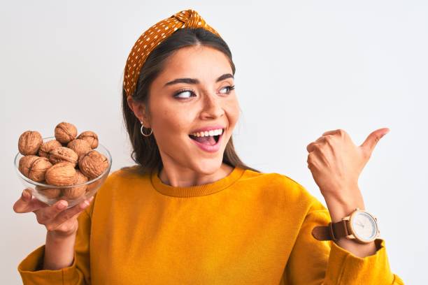 istock Young beautiful woman holding bowl with walnuts standing over isolated white background pointing and showing with thumb up to the side with happy face smiling 1175206862