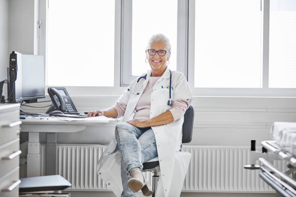 Portrait of smiling senior female doctor at desk Smiling senior female doctor sitting at desk. Portrait of confident healthcare worker is at hospital. She is in lab coat. legs crossed at ankle stock pictures, royalty-free photos & images