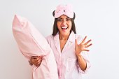 Beautiful woman wearing sleep mask pajama holding pillow over isolated white background very happy and excited, winner expression celebrating victory screaming with big smile and raised hands