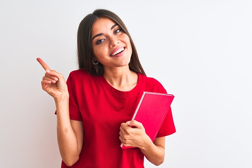 Young beautiful student woman holding red book standing over isolated white background very happy pointing with hand and finger to the side