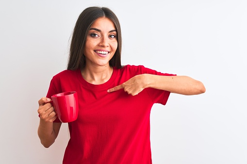 Young beautiful woman drinking red cup of coffee standing over isolated white background with surprise face pointing finger to himself