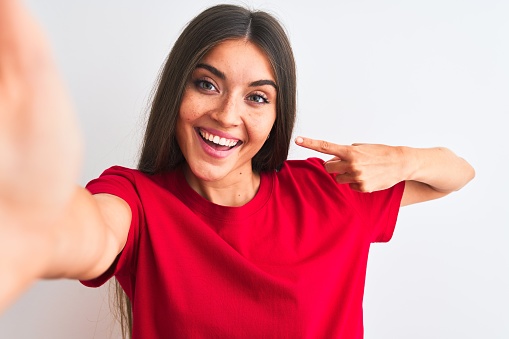 Beautiful woman wearing red t-shirt make selfie by camera over isolated white background very happy pointing with hand and finger