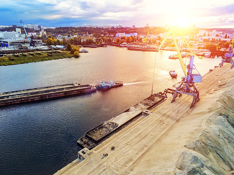 Aerial view of heavy crane loading bulk goods at Dnieper river cargo port terminal in Kiev at evening sunset time. tugboat pushes barge with sand after loading. Industrial inland navigation