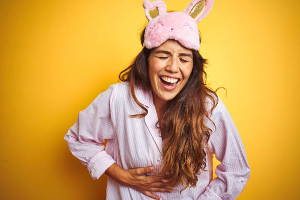 Young woman wearing pajama and sleep mask standing over yellow isolated background smiling and laughing hard out loud because funny crazy joke with hands on body. Young woman wearing pajama and sleep mask standing over yellow isolated background smiling and laughing hard out loud because funny crazy joke with hands on body. people laughing hard stock pictures, royalty-free photos & images