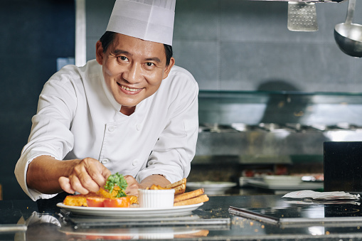 Successful Asian mature chef smiling at camera while decorating prepared dish with greens in the kitchen