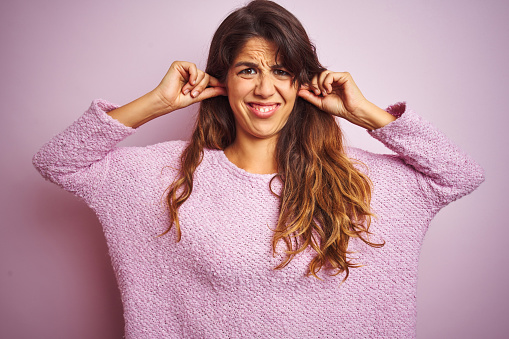 Young beautiful woman wearing sweater standing over pink isolated background covering ears with fingers with annoyed expression for the noise of loud music. Deaf concept.