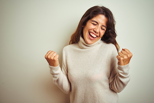 Young beautiful woman wearing winter sweater standing over white isolated background very happy and excited doing winner gesture with arms raised, smiling and screaming for success. Celebration concept.