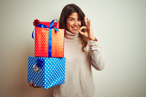Young beautiful woman holding birthday gifts standing over isolated white background doing ok sign with fingers, excellent symbol