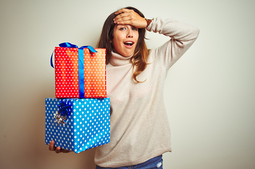 Young beautiful woman holding birthday gifts standing over isolated white background stressed with hand on head, shocked with shame and surprise face, angry and frustrated. Fear and upset for mistake.