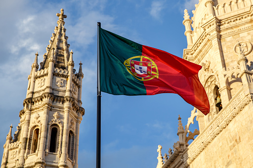 Portuguese flag waving in front of a blue sky and monastery.