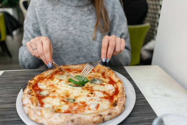 istock Woman eats with knife and fork a pizza Margherita with mozzarella tomatoes and basil. Neapolitan pizza from wood-burning stove. lunch in an Italian restaurant. Table near to a large window. 1175230997