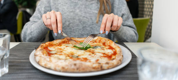 istock Woman eats with knife and fork a pizza Margherita with mozzarella tomatoes and basil. Neapolitan pizza from wood-burning stove. lunch in an Italian restaurant. Table near to a large window. 1175231052