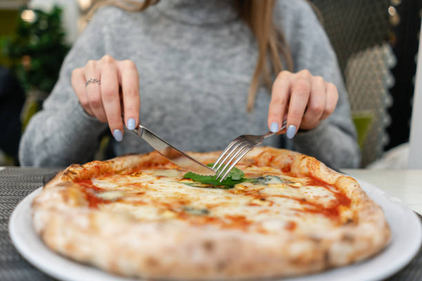 istock Woman eats with knife and fork a pizza Margherita with mozzarella tomatoes and basil. Neapolitan pizza from wood-burning stove. lunch in an Italian restaurant. Table near to a large window. 1175231073