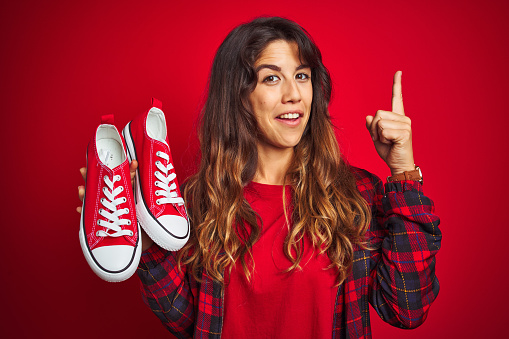 Young beautiful woman holding sneakers standing over red isolated background surprised with an idea or question pointing finger with happy face, number one
