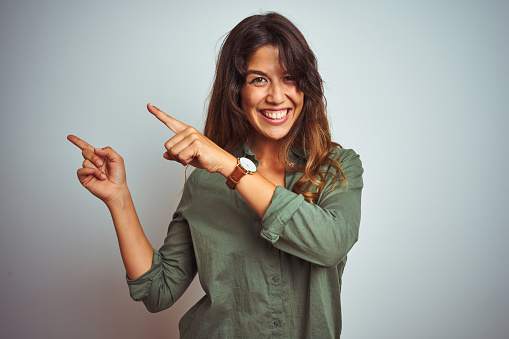 Young beautiful woman wearing green shirt standing over grey isolated background smiling and looking at the camera pointing with two hands and fingers to the side.