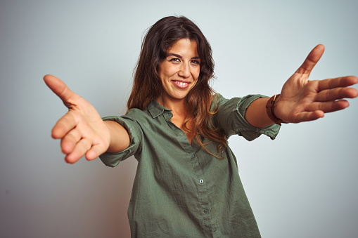Young beautiful woman wearing green shirt standing over grey isolated background looking at the camera smiling with open arms for hug. Cheerful expression embracing happiness.